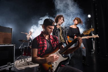 KYIV, UKRAINE - AUGUST 25, 2020: Rock band musician playing bass guitar sitting near curly vocalist and guitarist with smoke and female drummer on background clipart
