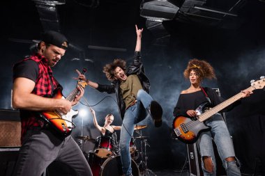 KYIV, UKRAINE - AUGUST 25, 2020: Vocalist with microphone screaming while jumping up near guitarists with backlit and female drummer on background clipart