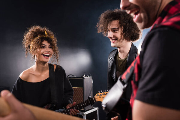 Smiling musician standing near curly woman with bass guitar with blurred guitarist on foreground