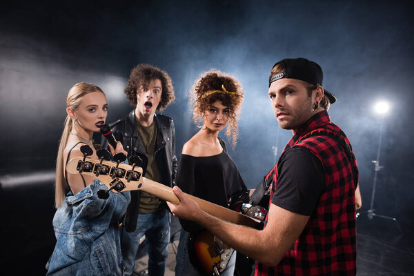 KYIV, UKRAINE - AUGUST 25, 2020: Rock band musicians with electric guitars and microphone looking at camera with backlit on black
