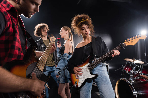 KYIV, UKRAINE - AUGUST 25, 2020: Guitarists playing electric guitars near talking rock band musicians with backlit and drum kit on black background