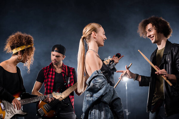 KYIV, UKRAINE - AUGUST 25, 2020: blonde vocalist talking with drummer standing near musicians playing electric guitars with backlit on black background