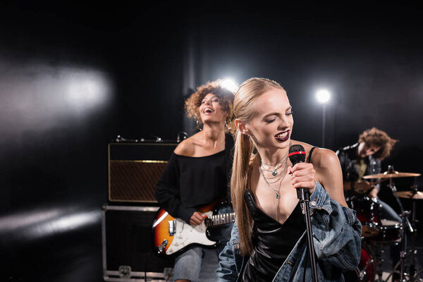 KYIV, UKRAINE - AUGUST 25, 2020: Blonde woman singing with microphone standing near guitarist with backlit and blurred drummer on background