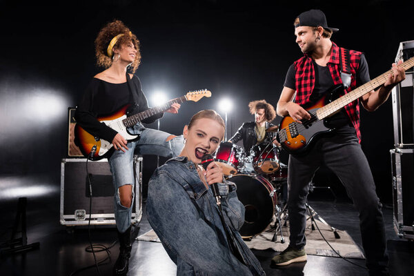 KYIV, UKRAINE - AUGUST 25, 2020: Female vocalist singing in microphone while sitting near guitarists with backlit and blurred drummer on background