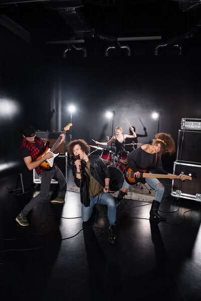 KYIV, UKRAINE - AUGUST 25, 2020: Rock band vocalist sitting on knee and shouting in microphone near guitarists with backlit and female drummer on black