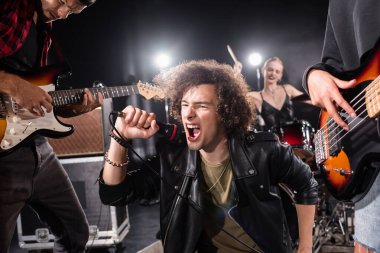 KYIV, UKRAINE - AUGUST 25, 2020: Curly vocalist shouting in microphone while sitting near guitarists with backlit and blurred female drummer on background clipart