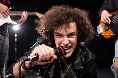 KYIV, UKRAINE - 25 AĞUSTOS 2020: Mikrofonlu kızgın vokalist kameraya arka ışıklandırılmış ve bulanık gitaristlerle bakıyor