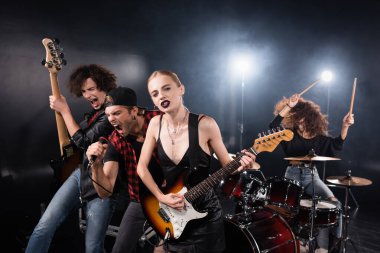KYIV, UKRAINE - AUGUST 25, 2020: Blonde woman looking at camera while playing electric guitar standing near musicians shouting and drummer on black clipart