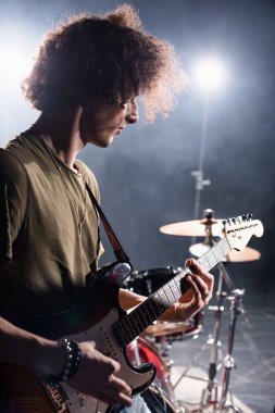 KYIV, UKRAINE - 25 AĞUSTOS 2020: Kıvırcık müzisyen arka planda bulanık arka planda davul setiyle birlikte bas gitar çalıyor