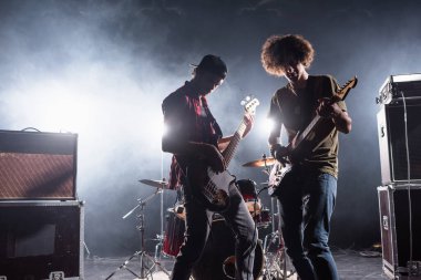 KYIV, UKRAINE - AUGUST 25, 2020: Rock band musicians playing bass guitars near drum kit and combo amplifiers with smoke and backlit on background clipart