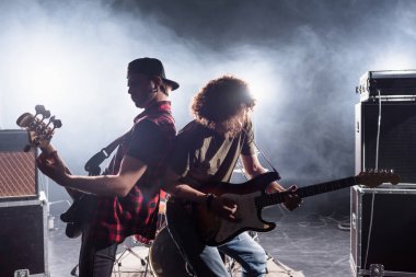 KYIV, UKRAINE - AUGUST 25, 2020: Rock band musicians standing back to back while playing electric guitars near combo amplifiers with smoke on background clipart