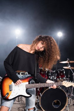 KYIV, UKRAINE - 25 AĞUSTOS 2020: Arka planda arka planda ışıklandırılmış davul setinin yanında eğilen ve bas gitar çalan kadın müzisyen