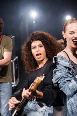 KYIV, UKRAINE - 25 AĞUSTOS 2020: Ağzı açık, elektro gitar çalan kıvırcık bayan müzisyen arka planı bulanık olan vokalistin yanında duruyor.