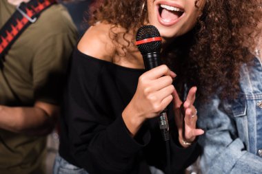 Cropped view of curly female vocalist with microphone singing near rock band musicians on blurred background clipart