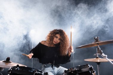 Female musician with drumsticks playing on drum kit while looking at camera with smoke on background clipart