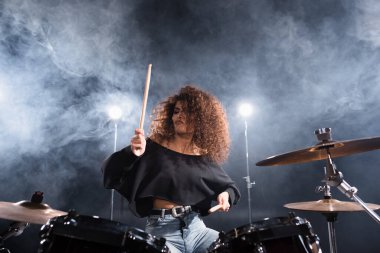 Curly woman with drumsticks playing on drum kit with smoke and backlit on background clipart