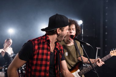 KYIV, UKRAINE - AUGUST 25, 2020: Rock band musicians shouting in microphone with blurred drummer on background clipart