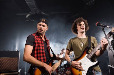 KYIV, UKRAINE - 25 Ağustos 2020: Bas gitar çalan müzisyenler mikrofon rafının yanında duruyor ve arka planda bulanık baterist ile karışık amplifikatörler