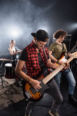 KYIV, UKRAINE - AUGUST 25, 2020: Rock band musicians playing electric guitars near drum kit with blurred woman on background clipart