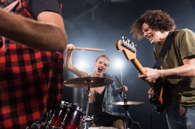 KYIV, UKRAINE - AUGUST 25, 2020: Blonde woman with drumsticks screaming while sitting at drum kit near guitarist with backlit and blurred man on foreground clipart