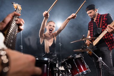 KYIV, UKRAINE - AUGUST 25, 2020: Blonde woman with outstretched hands screaming while sitting at drum kit near guitarist with backlit on blurred foreground clipart