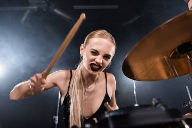 Blonde woman with drumstick winking and playing drum plate on blurred foreground clipart
