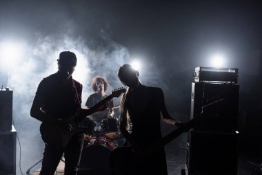 KYIV, UKRAINE - AUGUST 25, 2020: Rock band guitarists standing near combo amplifiers and drummer sitting at drum kit with backlit and smoke on background clipart