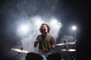 Excited curly drummer with open mouth sitting at drum kit with smoke and backlit on black  clipart