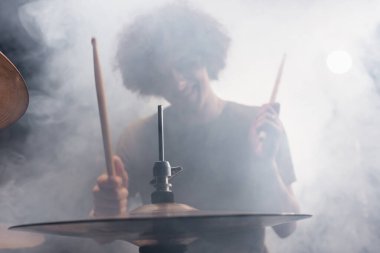 Smiling curly drummer with drumsticks playing drums in smoke clipart