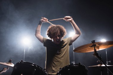 Curly musician shouting, while holding drumsticks and sitting at drum kit with backlit on background clipart