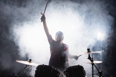 Drummer with hand in air, holding drumsticks while sitting at drum kit with smoke on black  clipart