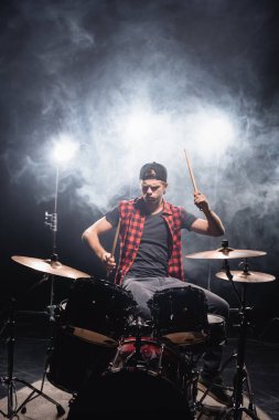 Serious musician with drumsticks playing drums with smoke and backlit on black  clipart