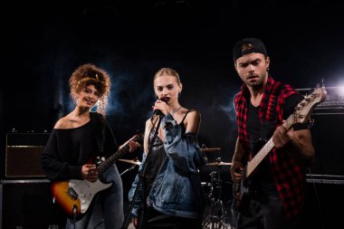 KYIV, UKRAINE - AUGUST 25, 2020: Female singer of rock band standing near musicians with electric guitars on black  clipart
