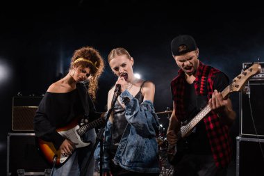 KYIV, UKRAINE - AUGUST 25, 2020: Blonde vocalist of rock band singing near musicians with bass guitars with backlit on black  clipart