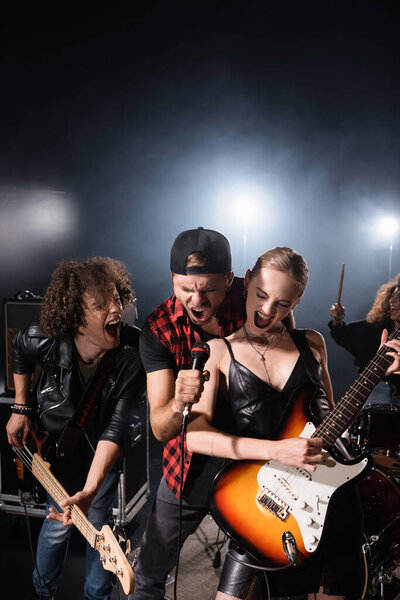 KYIV, UKRAINE - AUGUST 25, 2020: Vocalist shouting in microphone while standing near rock band guitarists with woman and backlit on black 