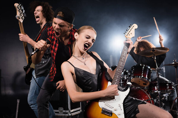 KYIV, UKRAINE - AUGUST 25, 2020: Blonde woman playing electric guitar while shouting with vocalist and guitarist with female drummer on blurred background