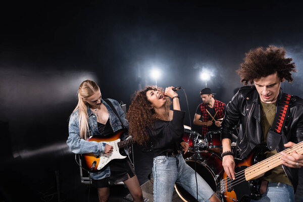 KYIV, UKRAINE - AUGUST 25, 2020:  Curly vocalist of rock band singing standing near musicians playing on drums and electric guitars with backlit on black