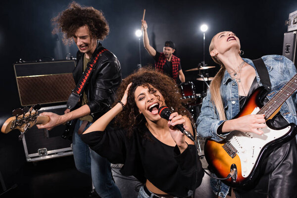 KYIV, UKRAINE - AUGUST 25, 2020:  Female singer of rock band with microphone singing near guitarists with blurred drummer on background