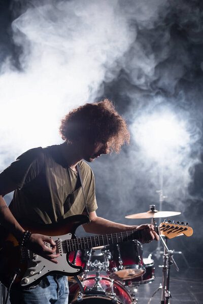KYIV, UKRAINE - AUGUST 25, 2020: Rock band musician playing guitar with pick near drum kit with smoke on background