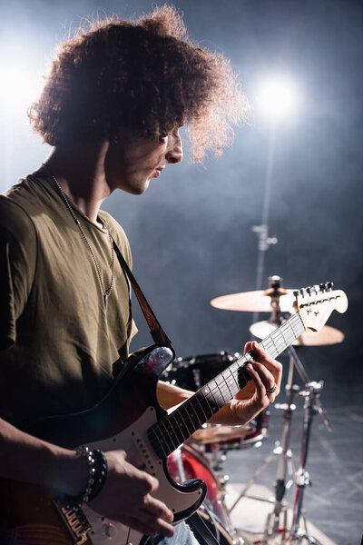 KYIV, UKRAINE - AUGUST 25, 2020: Curly musician playing bass guitar while standing near drum kit with backlit on blurred background