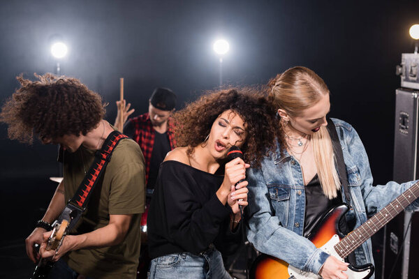 KYIV, UKRAINE - AUGUST 25, 2020: Curly vocalist with closed eyes singing near guitarists with blurred drummer on background