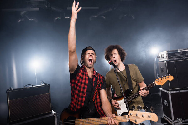KYIV, UKRAINE - AUGUST 25, 2020: Rock band musicians shouting while holding guitars near microphone rack and combo amplifiers with backlit on background