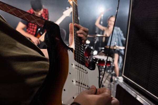 KYIV, UKRAINE - AUGUST 25, 2020: Close up view of man playing electric guitar near combo amplifiers with blurred drummer and guitarist on background