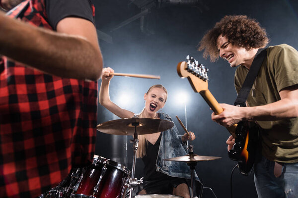 KYIV, UKRAINE - AUGUST 25, 2020: Blonde woman with drumsticks screaming while sitting at drum kit near guitarist with backlit and blurred man on foreground