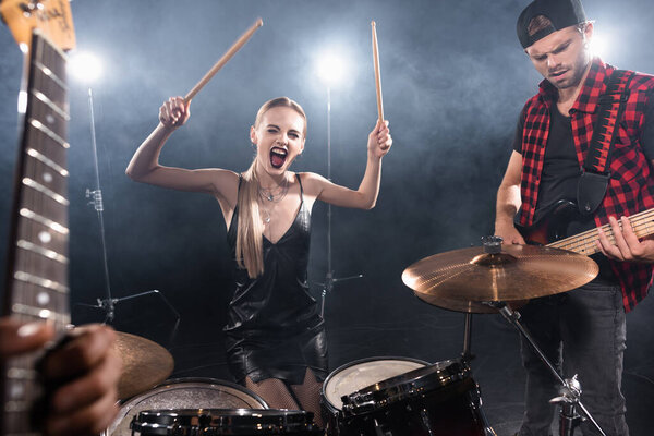 KYIV, UKRAINE - AUGUST 25, 2020: Blonde woman with drumsticks shouting while sitting at drum kit near guitarist with backlit and blurred guitar on foreground