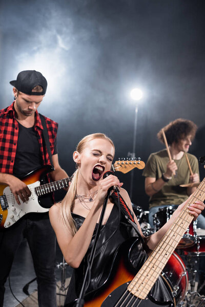 KYIV, UKRAINE - AUGUST 25, 2020: Female vocalist of rock band shouting in microphone near guitarist with backlit and blurred drummer on background