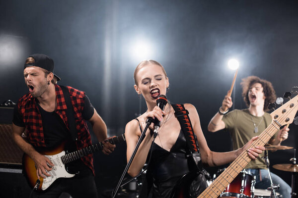 KYIV, UKRAINE - AUGUST 25, 2020: Blonde vocalist with electric guitar singing in microphone near guitarist and drummer with backlit on blurred background