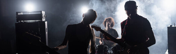 KYIV, UKRAINE - AUGUST 25, 2020: Rock band guitarists standing near combo amplifiers and drummer sitting at drum kit with backlit and smoke on background, banner