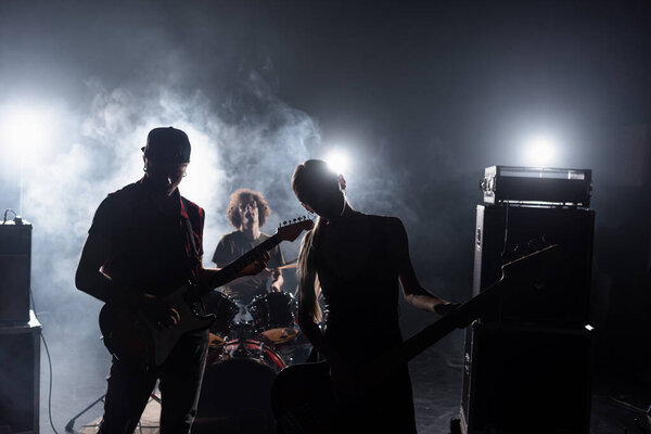 KYIV, UKRAINE - AUGUST 25, 2020: Rock band guitarists standing near combo amplifiers and drummer sitting at drum kit with backlit and smoke on background