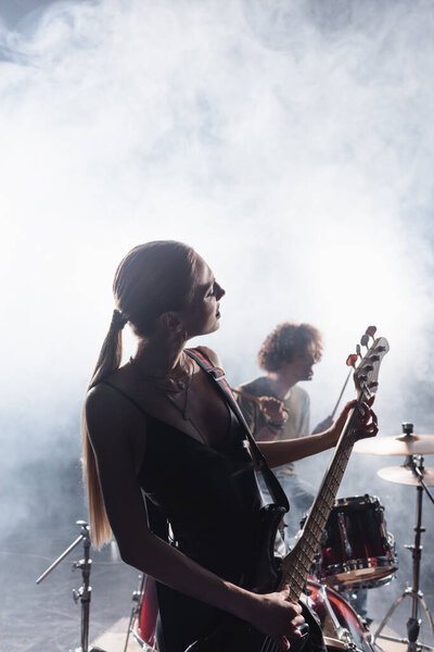 KYIV, UKRAINE - AUGUST 25, 2020: Woman playing electric guitar with smoke and blurred drummer with drum kit on background